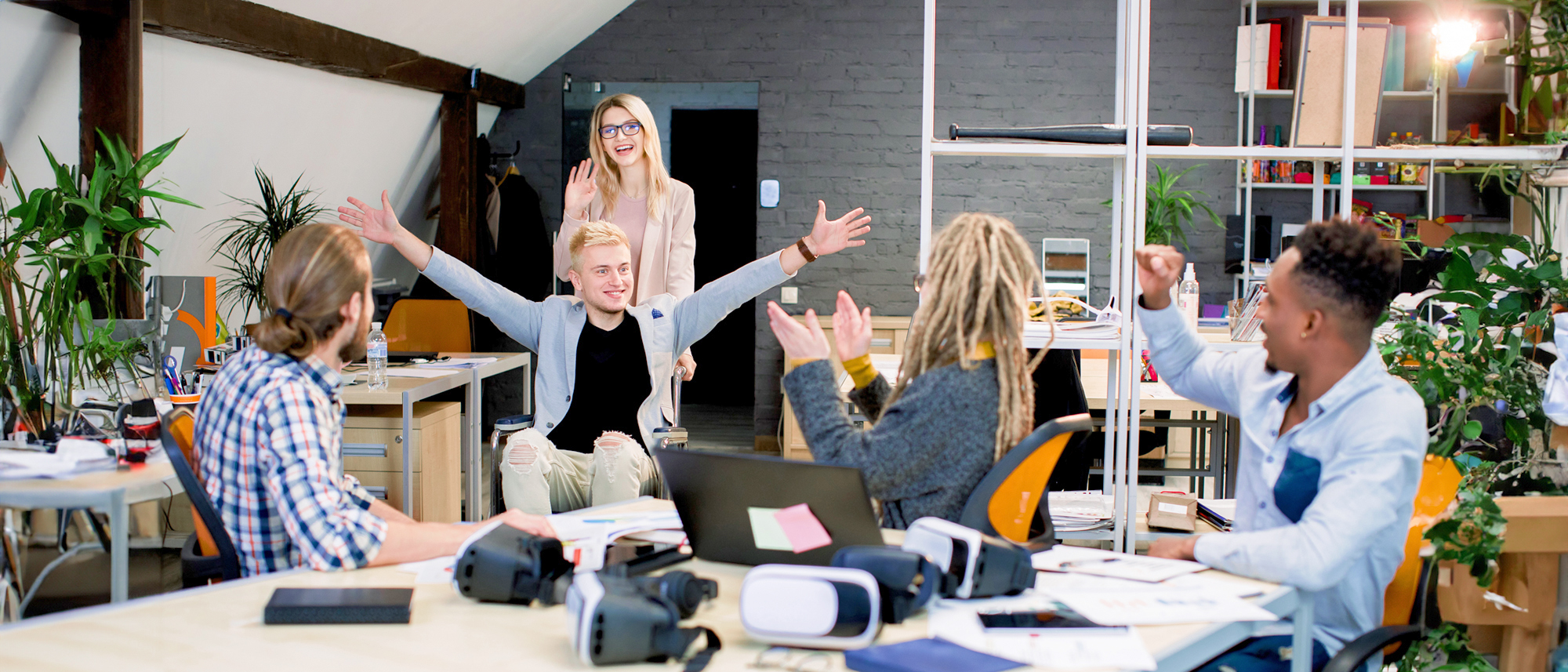 Eine Gruppe von Menschen sitzt an einem Tisch, mit erhobenen Armen in einem Büro. Sie scheinen fröhlich und engagiert zu sein. Eine Person aus dem Team sitzt im Rollstuhl.